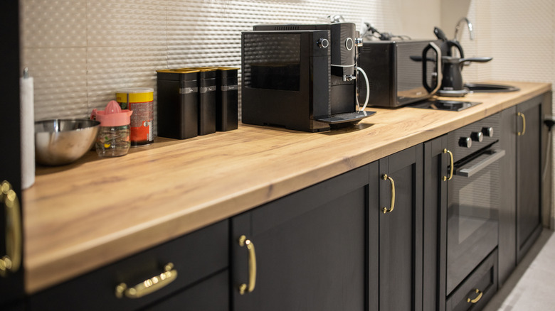 A closeup photo of a kitchen, decked with black cabinets, a wooden worktop, white backsplash, and black appliances