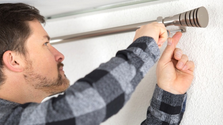 man hand-screwing curtain rod into white textured wall near ceiling