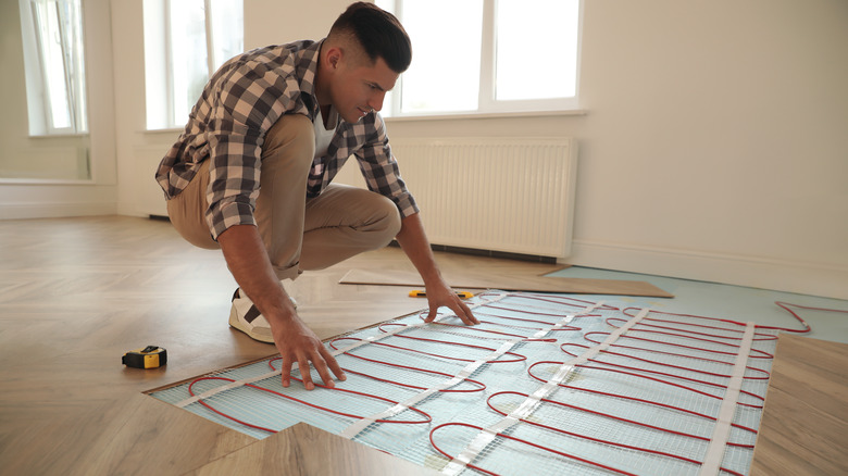 A man crouches to inspect radiant heat components under a wooden floor