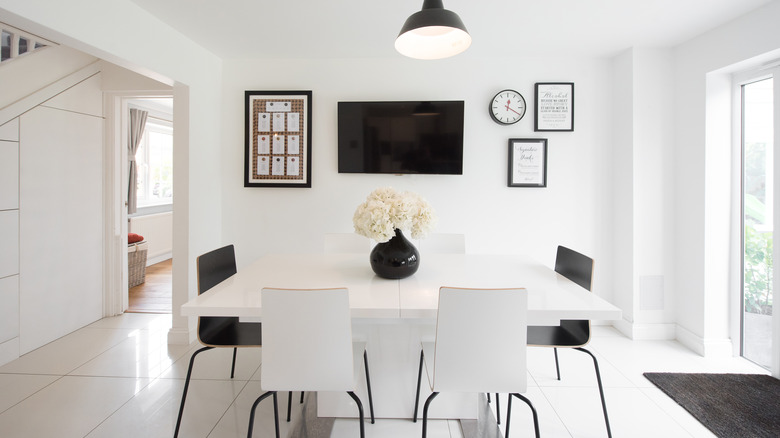 A room with white walls, a white dining set, a television on the wall, a wall clock and frames