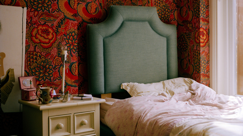 A colorful bedroom is designed with patterned wallpaper, upholstered headboard, and trinkets on nightstand