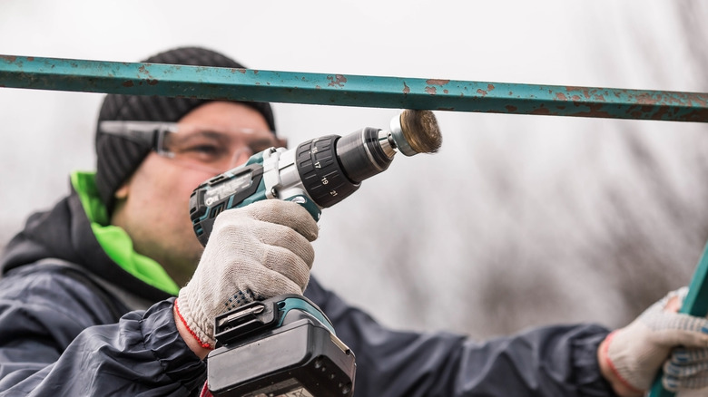 A man uses a drill with a steel wool attachment to scrape off rust from a bar