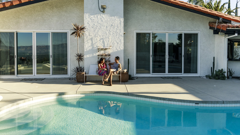 Two people sit by a pool with a concrete pool deck.