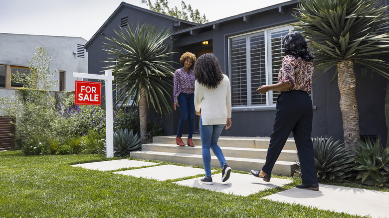 Three women standing in front of an upscale house with a for sale sign