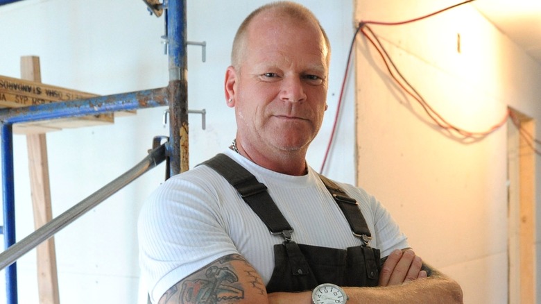 Mike Holmes in overalls in unfinished room with hanging wires and scaffold