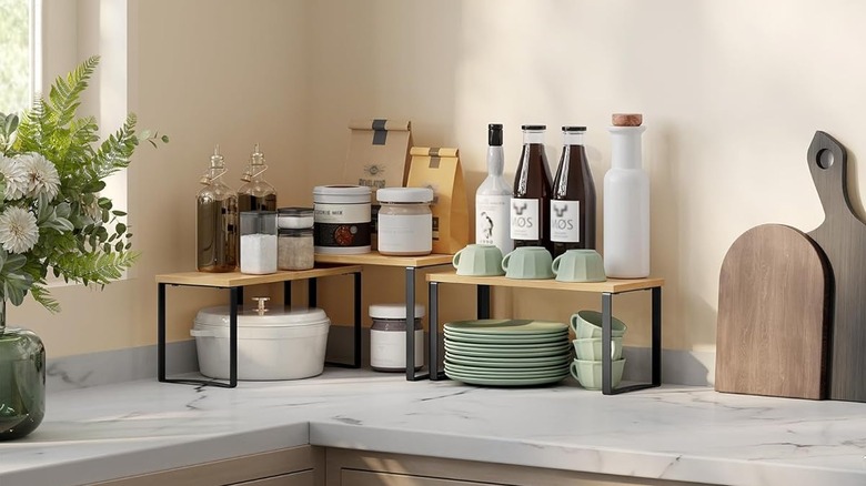 A kitchen countertop corner is neatly organized thanks to the use of three shelf risers storing various containers on top and plates, mugs, and a pot beneath.