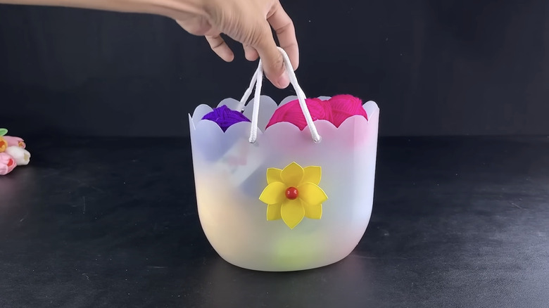 A hand holds up a DIY yarn basket made from a plastic bottle.