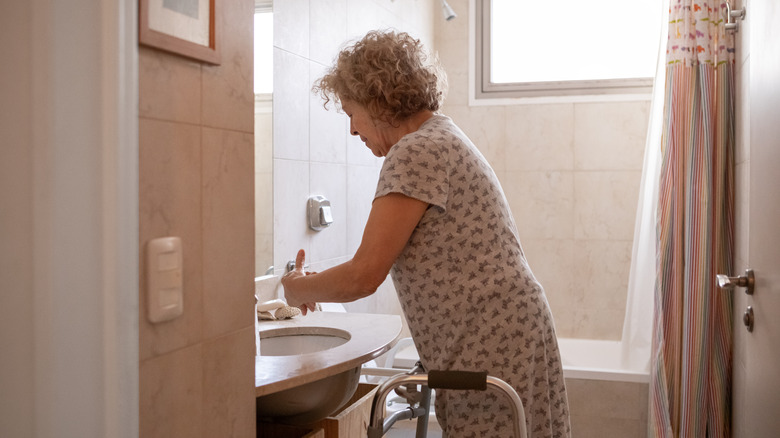 Older woman washes her hands with a walker at her side in a bathroom with a shower-tub combo