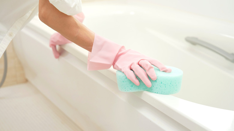 Woman cleans white bathtub with a green sponge while wearing pink gloves