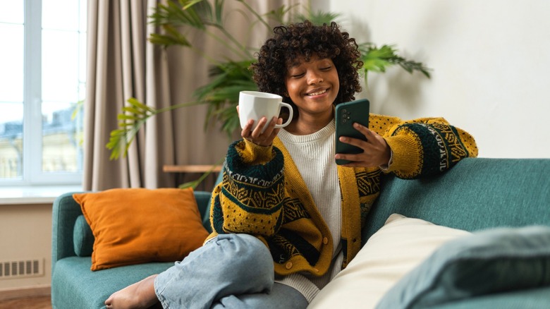 A person on green couch holding phone and coffee mug