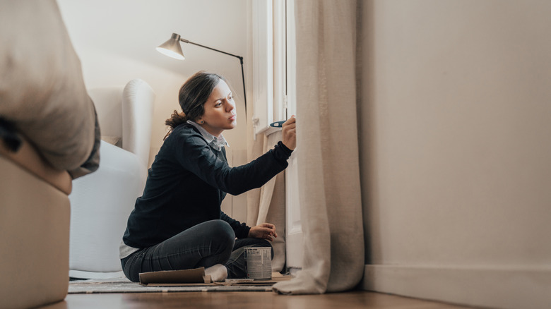 Woman sitting cross-legged on the floor painting window trim in beige living room