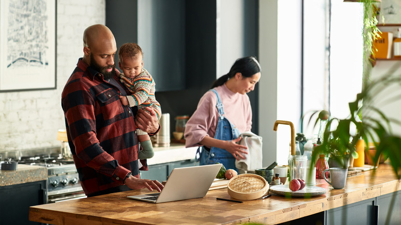 A man holding a baby stands beside a woman in a bright, open kitchen.