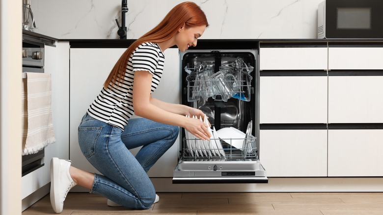 Woman kneeling beside open dishwasher and removing plate from rack