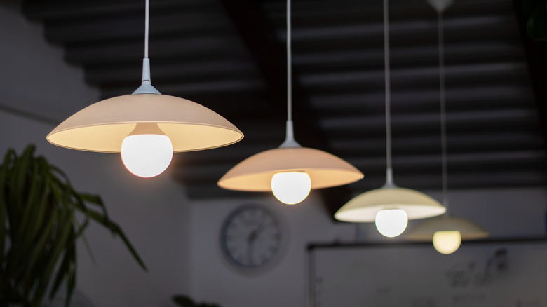 A series of simple, uniform hanging light fixtures with a clock and plant in background