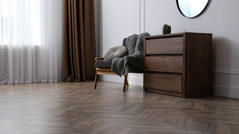 Dim interior room with linoleum parquet flooring, dresser, and chair beside sunny window