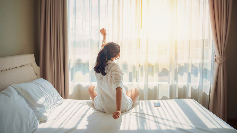 A woman stretches in bed in front of a sunny window