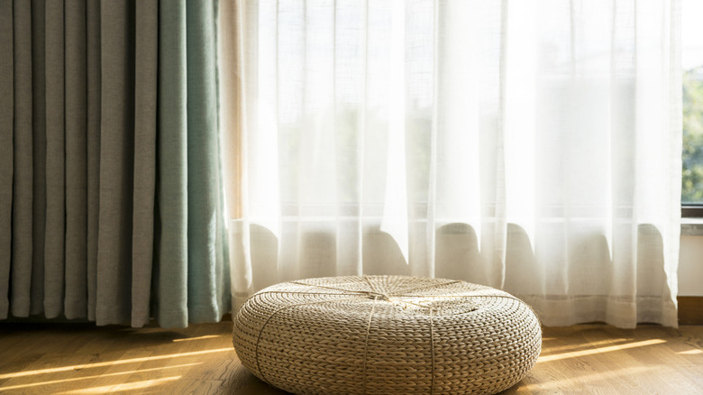 Layers of curtains in a window are behind a seat cushion on the floor