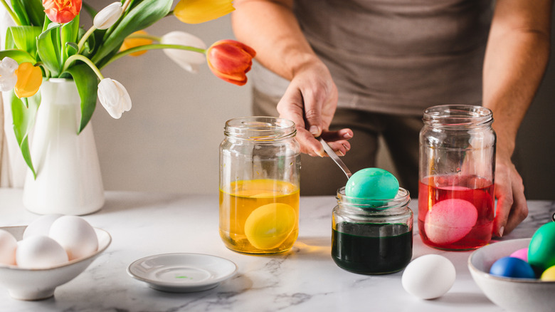 A person dyes Easter eggs in three clear glass jars of colorful dye solution