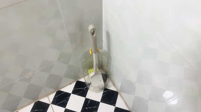 Plastic scrub brush inside of a plastic jug in the corner of a tile floor shower