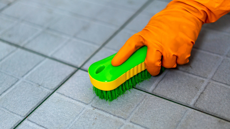 Orange gloved hand holds green brush and scrubs grey tile