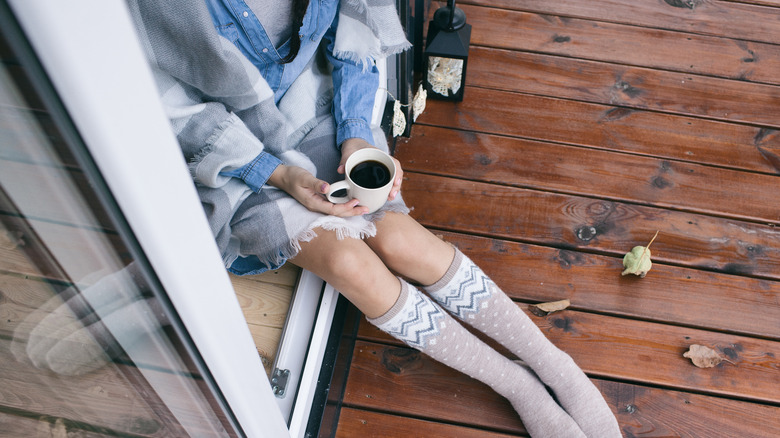 Woman wrapped in a blanket and holding a mug sits in doorway on wooden deck