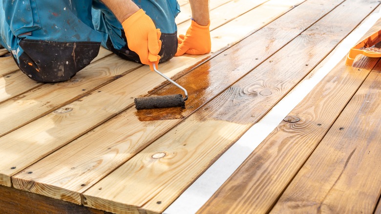 Kneeling person wearing gloves uses a roller to apply sealant to wood deck