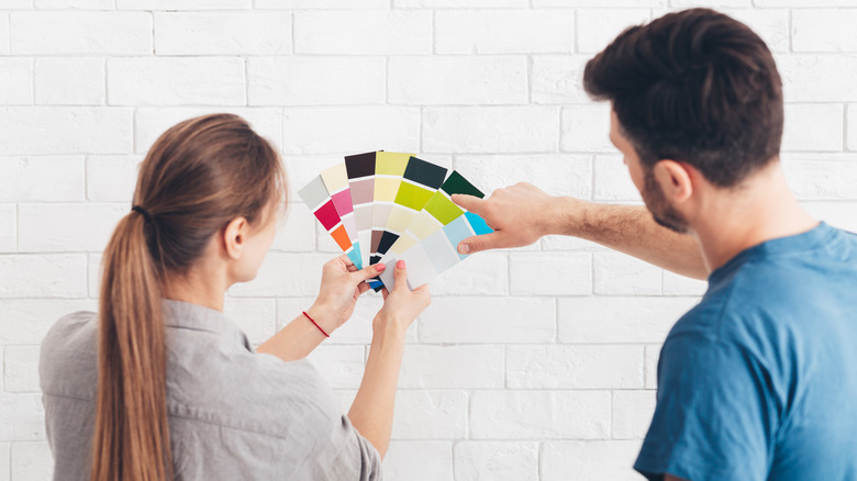 A couple holds a color paint palette while facing a white wall.