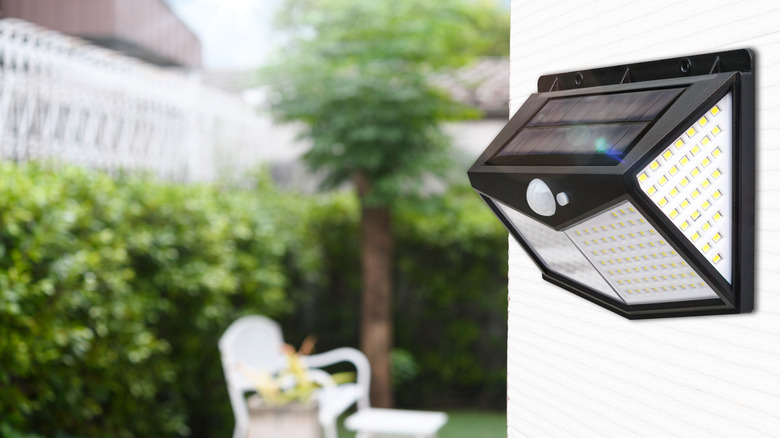 solar powered motion detector light installed on exterior wall with yard chair and greenery in the background