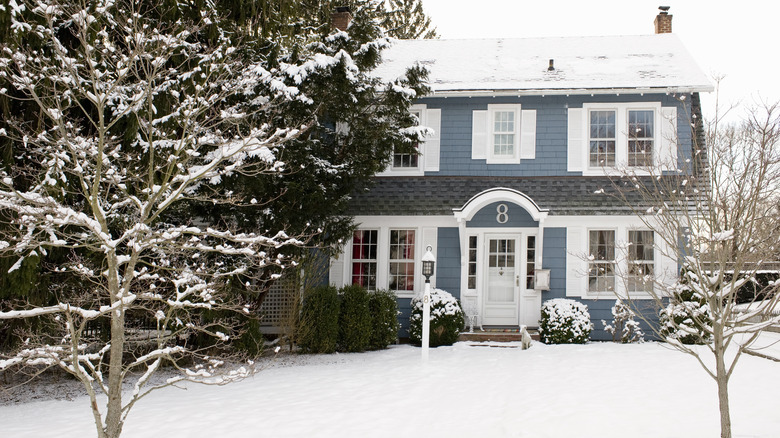 A blue multi-level house with a snow-covered yard, trees, and roof