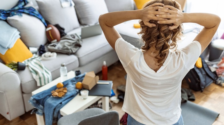 A back view of a woman holding her head while looking at a cluttered living room sofa