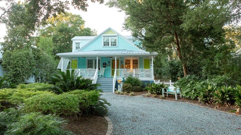 A gravel driveway surrounded by lush green trees and bushes leading up to a pale blue house