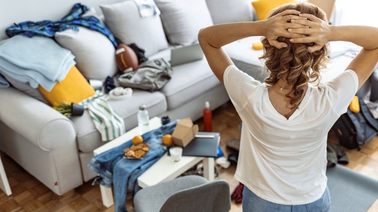 Woman in white tshirt and jeans clasping head starting at messy living room sofa and coffee table