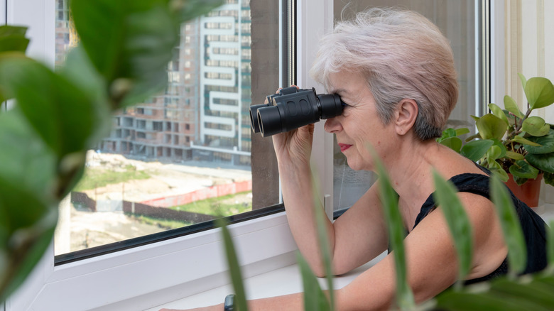 An elderly woman with binoculars spying out a window