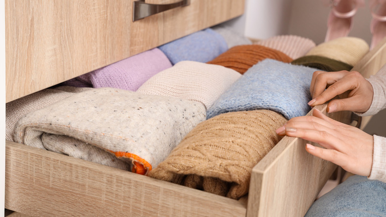 A person's hands are seen at the edge of a tidy, pale wood sweater drawer.