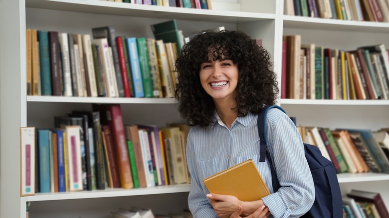 A woman holding a book next to a bookshelf.