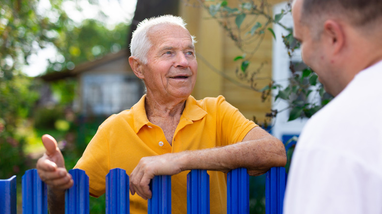 Elderly man talking to his neighbor over a blue fence.