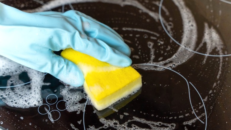 closeup of hand in blue rubber love cleaning glass stovetop with yellow sponge and soapy water