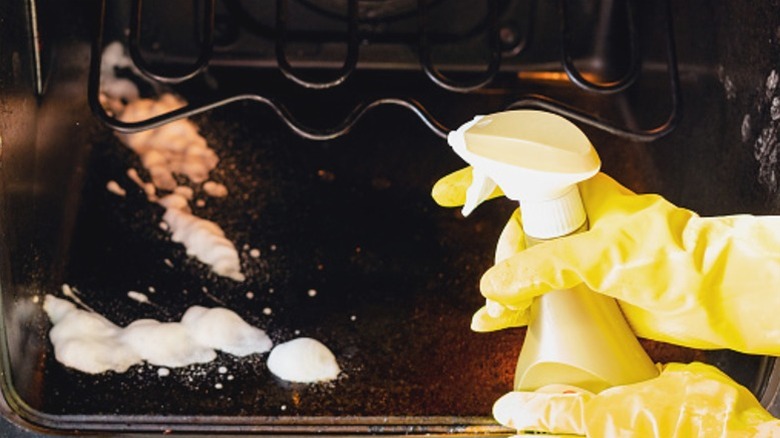 person in yellow rubber gloves spraying foam cleaner into dirty oven