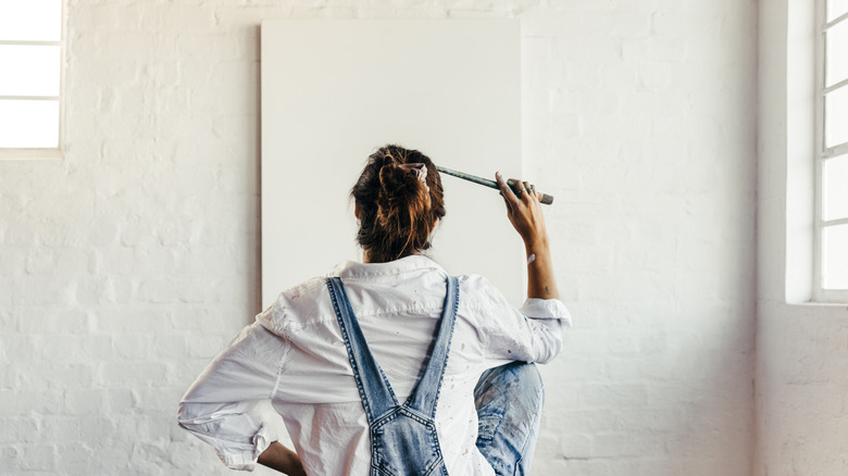 A woman wearing overalls stares at a blank canvas on a white wall while holding a paintbrush