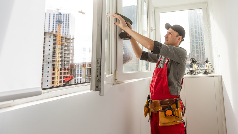 Young man wearing overalls sealing cracks between window and trim using waterproof silicone caulk on the balcony.