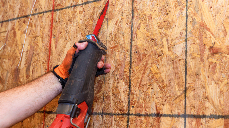 A hand using a reciprocating saw to cut through OSB