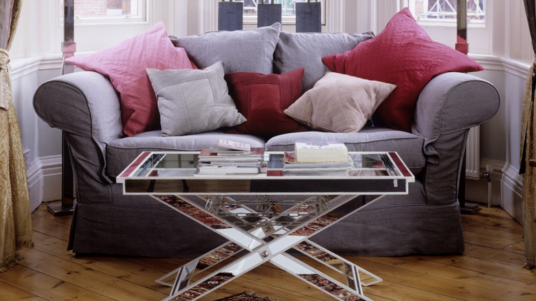 A mirrored coffee table is shown in front of a gray loveseat with red and cream pillows.