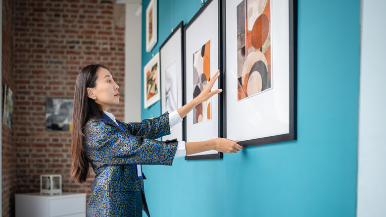 A woman arranges artwork in similar frames on a blue wall