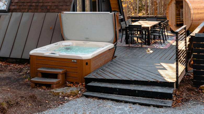 A hot tub sits next to a large wooden deck with a table and barrel sauna