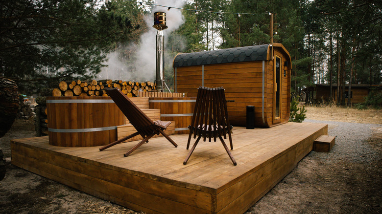 A cord of wood, two chairs, and a steaming sauna sit on a wooden platform deck