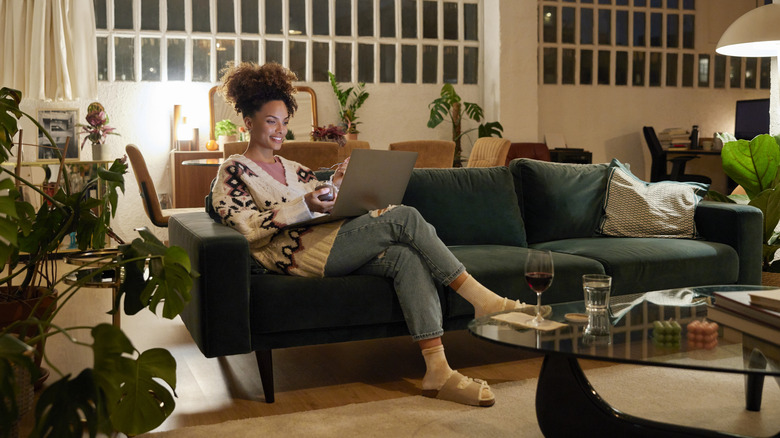 A woman sits on her couch at home while browsing her laptop and drinking wine.