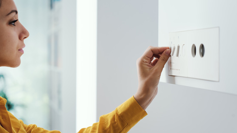 Woman in yellow shirt in a white room trying to turn on a boiler mounted to the wall