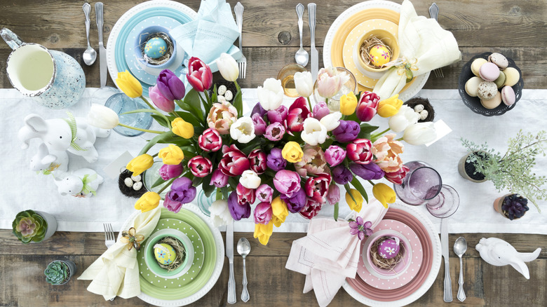 A colorful, narrow centerpiece sits on a wooden table with tulips and Easter-themed decor and dishes.