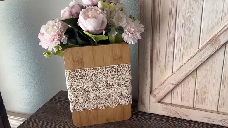 A natural wood rectangle vase with lace details holds a beautiful assortment of light pink faux flowers.