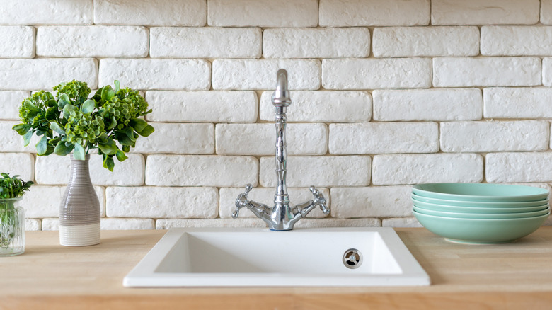 Kitchen sink in front of brick-like backsplash, topped with white cupboards.